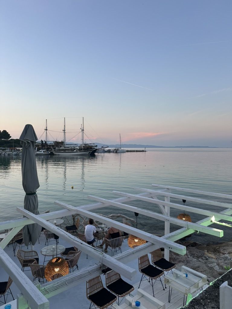 The terrace of On the Rocks at dusk, with the harbour and tall ships of Ormos Panagias behind
